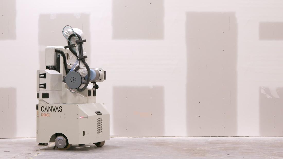 Canvas 1200CX drywall finishing robot. A white mobile base carries an arm with a sanding head, covered in a protective suit, before a wall with joint-compound patches.