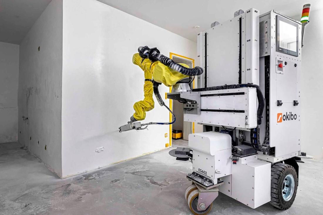 Okibo EG602 finishing an interior wall. A mobile robot with an arm in a yellow protective cover works on a white wall in an unfinished room; the unit is labeled EG602.