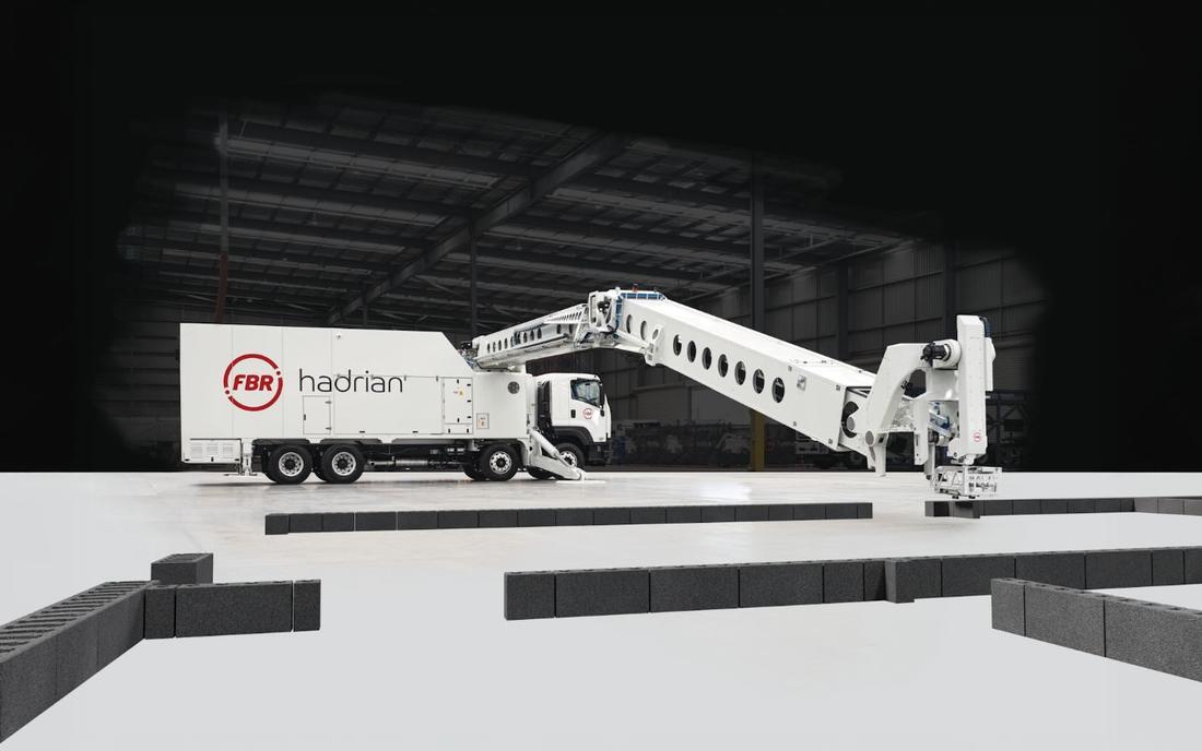 Hadrian brick-laying truck in a large hall. A white truck with a long telescopic boom extended over dark block courses on a floor inside a large hall; the image looks staged.