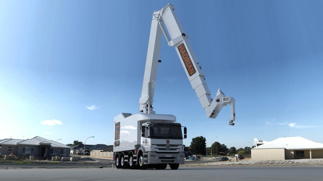 Hadrian X render on a residential street. Where: Residential street (location not stated). A white truck with its boom raised stands on a street with houses under construction behind it. It appears to be a CGI render composited onto a photo (2017); it does not show a real laying operation.