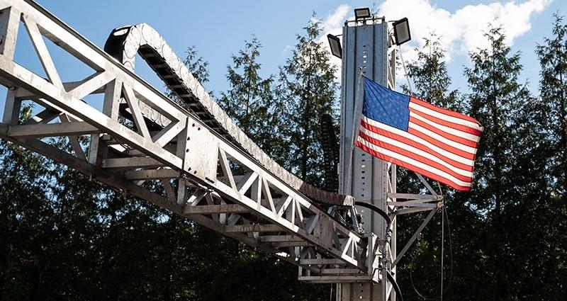 SQ4D ARCS gantry arm and mast. A truss gantry arm and mast of the ARCS printer rise against trees and sky; a US flag is mounted on the mast. Only hardware is visible; no printed structure appears in the frame.