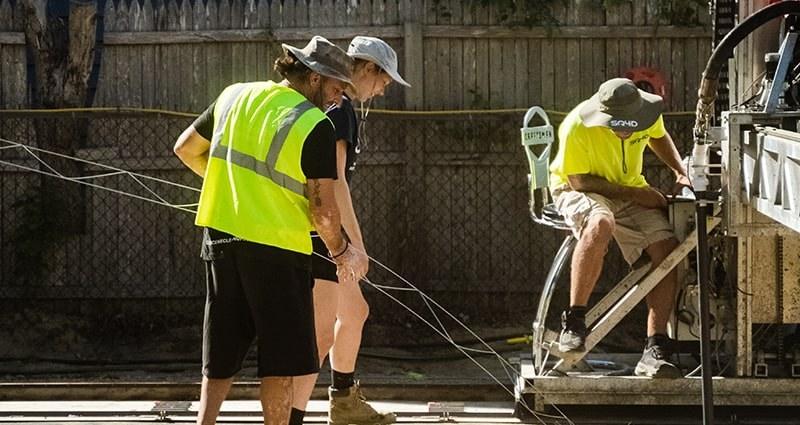 SQ4D crew beside the ARCS equipment. Three crew members in hi-vis stand beside printer equipment on a residential job site behind a wooden fence. The frame is tightly cropped and the printer shows only at the right edge.