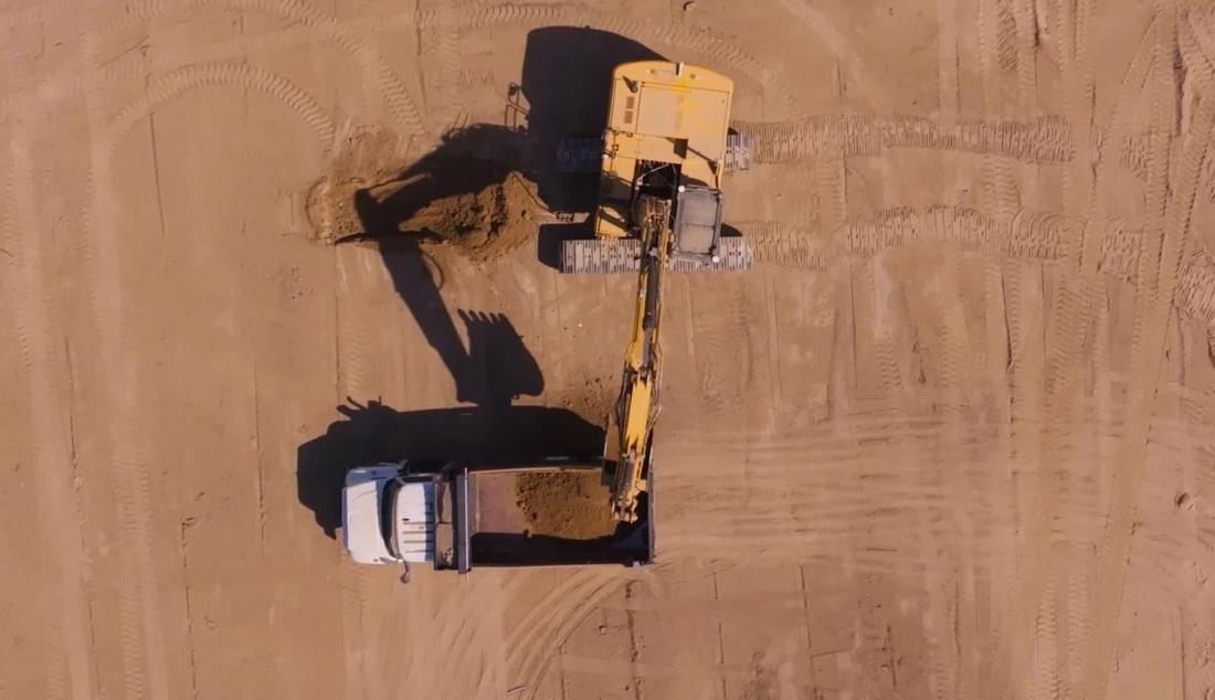 Excavator loading a dump truck, aerial view. A top-down view of a yellow tracked excavator loading soil into a white dump truck on bare, track-marked dirt. This is a frame from Bedrock's homepage; the autonomy retrofit itself is not visible.