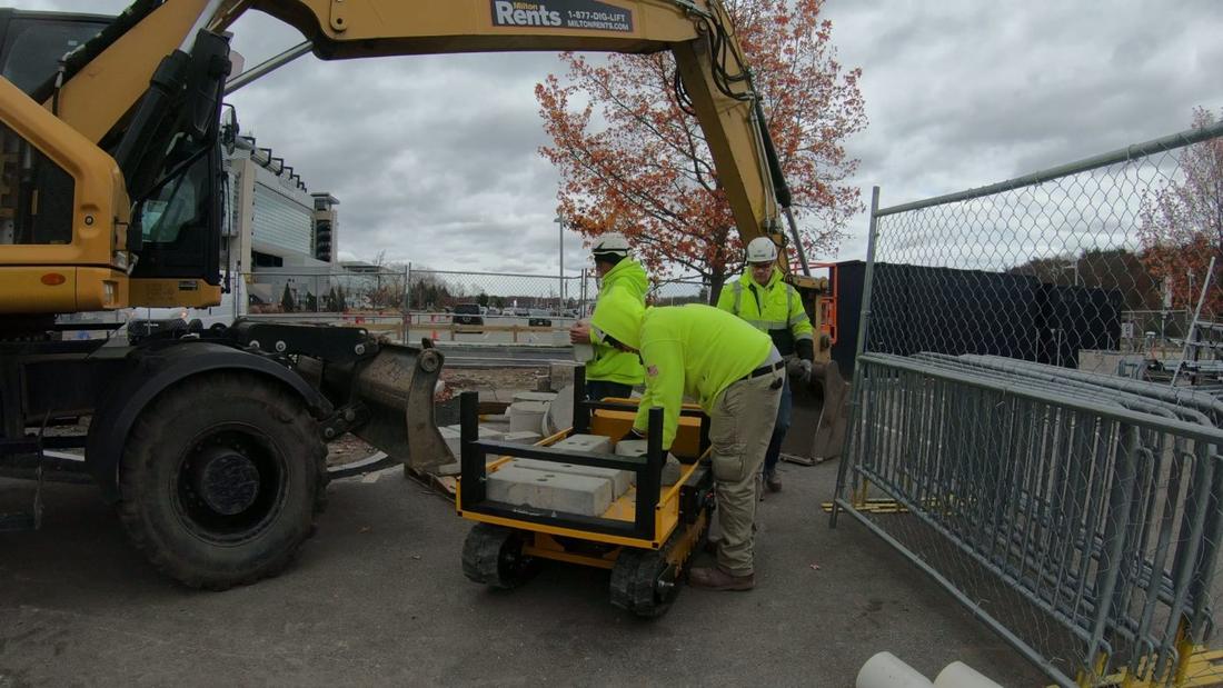 Tracked hauler cart loaded with pavers. Workers load concrete pavers onto a small yellow tracked hauler cart beside an excavator at a fenced construction site.